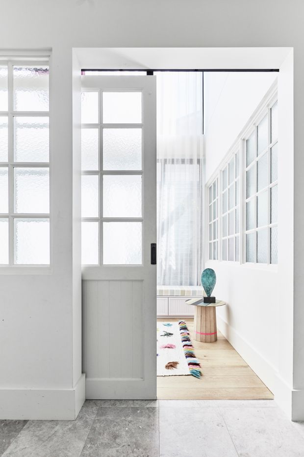 Bright, minimalist interior view through a partially open white glazed door into a light-filled room. The space features extensive white-framed multi-pane glazing, sheer curtains, pale timber flooring, white vertical wall panelling, a small cylindrical timber side table with blue decorative sculpture, and a colourful runner rug. Grey stone or concrete-look tiles are visible in the foreground hallway.