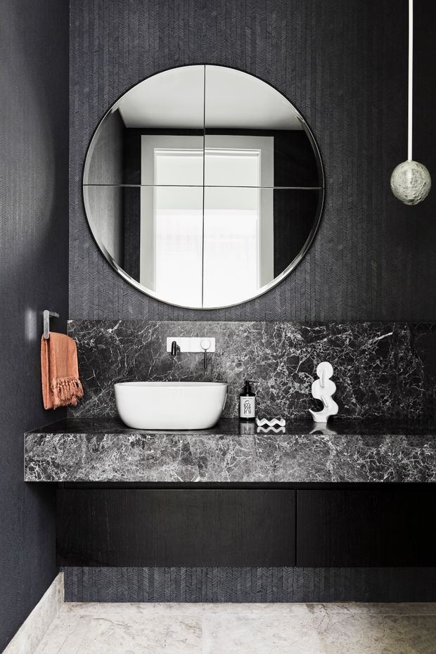A compact, dramatically styled bathroom vanity featuring a floating dark timber cabinet, white vessel basin, black marble benchtop and backsplash with prominent white veining, and a large round segmented mirror. The walls are clad in dark textured patterned wallpaper, with a sculptural pendant light and warm terracotta hand towel adding contrast.