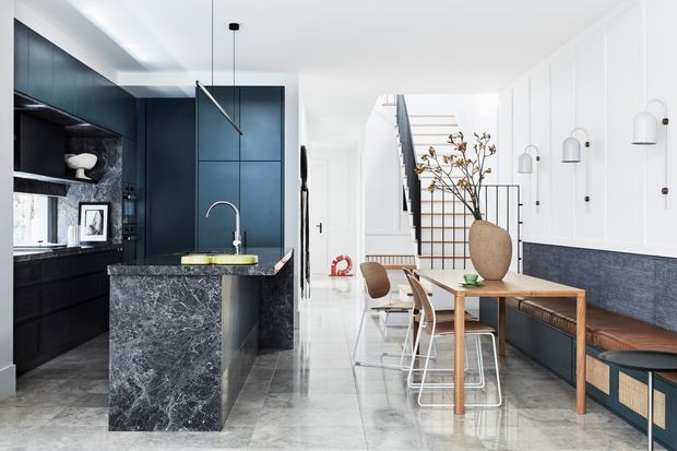 Open-plan contemporary kitchen and dining area with a dark blue-black kitchen installation, dramatic veined stone island, white paneled walls, polished light stone flooring, and a timber dining table with mixed seating. A stairway and bench seating are visible in the background, with sculptural pendant and wall lighting throughout.