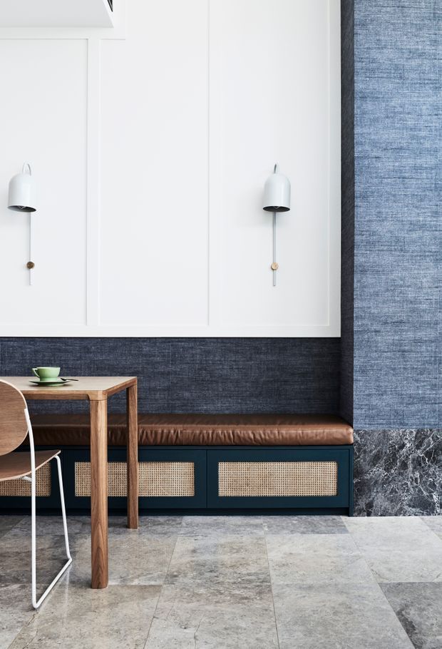 Refined contemporary dining or café-style interior featuring a white wall with geometric panel moulding and minimalist wall sconces, a dark blue textured wall treatment, built-in dark cabinetry with woven cane fronts, and a brown leather banquette. A simple timber dining table and chair sit on a large-format grey stone-look tiled floor, with a dark veined stone feature at the right.