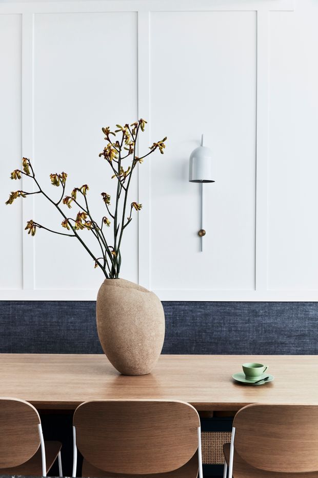 A refined dining area with a long light timber dining table and dark wood dining chairs, set against a white wall with decorative panel moulding. A sculptural neutral ceramic vase with delicate branches forms the centrepiece, while a minimalist wall sconce and small green cup add subtle detail. The composition is clean, balanced, and softly contemporary with muted natural tones.