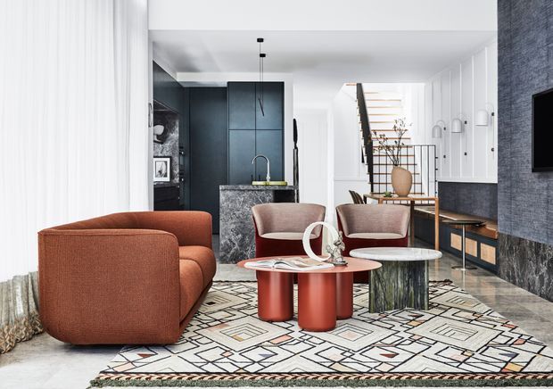 A bright, contemporary open-plan living room with a compact dark kitchen at the rear, sculptural lounge seating, red cylindrical coffee tables, a round stone-topped side table, and a large geometric patterned rug. White built-in cabinetry and a stairway with black railings line the right side, while textured dark wall finishes and marble-like stone surfaces add contrast.