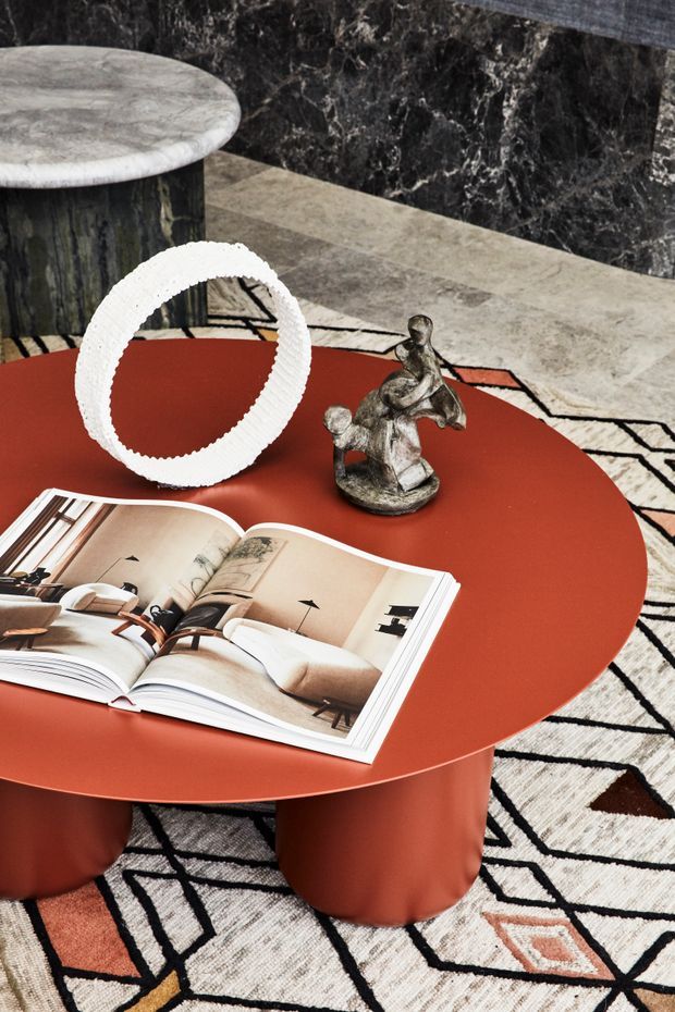 A styled living room vignette featuring a round terracotta-red coffee table with sculptural cylindrical legs, an open interior-design book, a white textured circular sculpture, and a small metallic figurine. The table sits on a bold geometric black, cream, terracotta, and grey rug, with a dark veined marble wall and a round marble-topped side table in the background.