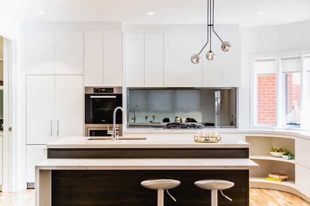 Bright contemporary kitchen with extensive white full-height cabinetry, integrated appliances, a large central island with a dark timber-look base and pale benchtop, a stainless steel sink and tap, and two sculptural stools. A reflective grey splashback, pendant lighting, and a curved window-seat/breakfast nook with open shelving complete the space.