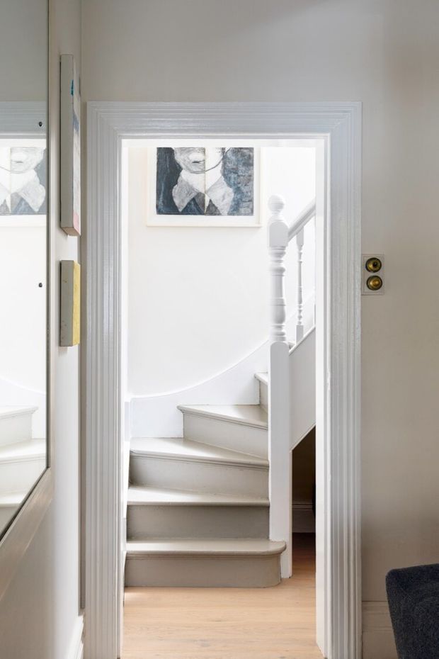 A bright, narrow interior view through a richly moulded white doorway toward a curved staircase. The stair has painted white risers and balustrade, pale timber flooring, light neutral walls, and framed monochrome artwork. A partial mirror and artwork are visible at left, with a small dark upholstered element at lower right. The space feels refined, airy, and traditional in its detailing.