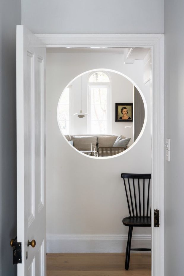Bright white traditional interior viewed through an open paneled door, featuring decorative mouldings, a circular interior window, pale timber flooring, and a black spindle-back chair. Through the circular opening, a light-filled sitting room with an upholstered sofa, arched window, pendant light, and framed artwork is visible.