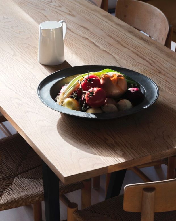 A sunlit dining table with a light timber top and dark angled legs, surrounded by wooden chairs with woven seats. A black shallow serving bowl filled with fruit and vegetables sits at the centre, alongside a white ceramic jug. The image has a warm, natural, minimalist dining setting.