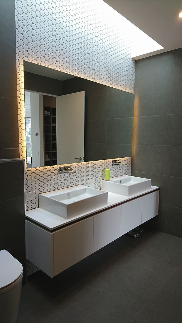 Contemporary bathroom featuring a floating double vanity with two white vessel basins, dark large-format wall and floor tiles, and a white hexagonal tile splashback extending around a large illuminated mirror. A skylight or large ceiling opening provides strong natural light, while concealed lighting adds a warm glow.