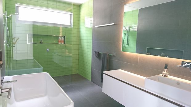 A contemporary bathroom with a freestanding white bathtub in the foreground, a glass-enclosed shower, bright green tiled walls, dark grey floor and feature wall tiles, and a long white vanity with integrated basins beneath a large illuminated mirror. The room is clean, bright, and strongly colour-blocked.