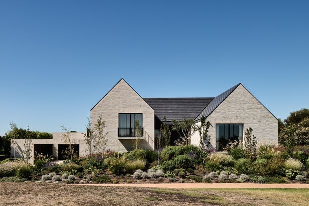Exterior view of a low-rise residential home composed of several steep-gabled volumes. The facade is clad in light textured masonry, with dark-framed windows and a dark pitched roof. A landscaped front garden with layered planting and a paved pedestrian path sits in front of the house under a clear blue sky.