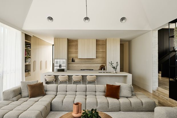Open-plan contemporary living area with a large modular sectional sofa in the foreground and a minimalist kitchen behind it. The kitchen features a long island with four upholstered stools, pale wood cabinetry, a wood-grain splashback, integrated appliances, and a light stone-coloured benchtop. Floor-to-ceiling sheer curtains line the left side, while a dark stairway or circulation zone is visible at right. The room has a bright, warm-neutral palette and clean built-in detailing.