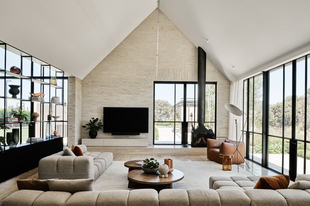 A bright, spacious open-plan living room with a dramatic steeply pitched ceiling and full-height glazing. Pale textured brick walls frame a central television and black freestanding fireplace, while a large curved sectional sofa, leather lounge chairs, round timber coffee tables, and open black metal shelving furnish the space. The room has strong indoor-outdoor visual connection and a refined contemporary feel.
