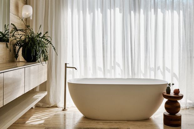 A serene, warmly lit contemporary bathroom featuring a freestanding oval bathtub positioned in front of full-height sheer curtains. A slim dark metal floor-mounted bath tap stands beside the tub, while a wall-mounted timber vanity with open shelving, pale cabinetry, a sculptural wall light, and lush potted greenery occupies the left side. The room has a light timber floor and a calm neutral palette with soft natural light.