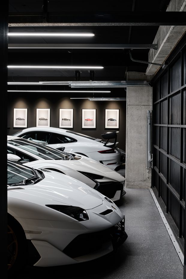 A dark, industrial-style luxury car showroom or private vehicle storage space. Several white high-performance sports cars are parked in rows beneath linear LED strip lighting. The space features exposed concrete columns and beams, black ceilings and structural framing, dark metal-framed glazing or garage doors, and a charcoal feature wall displaying framed automotive photographs.