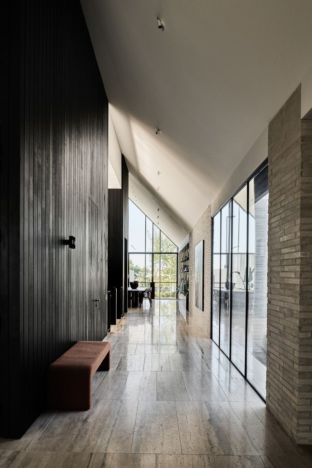 A long, dramatic interior corridor or gallery-like passage with a sloping white ceiling, tall black-framed glazing, and a polished pale stone floor. The left wall is clad in vertically ribbed dark timber or black panelling with a concealed door and a low upholstered bench. The right side combines exposed light brickwork with extensive glass partitions, revealing a bright adjoining space. Minimal recessed and surface lighting, strong linear perspective, and abundant daylight create a refined contemporary atmosphere.