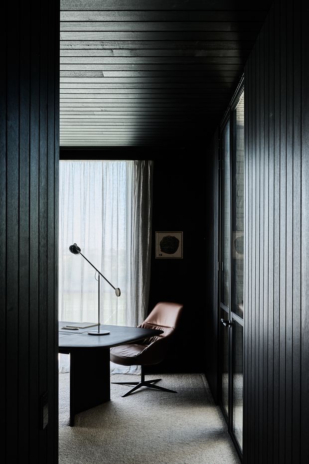 A narrow, darkly finished home office or study viewed through a doorway, with black vertical timber wall and ceiling panelling, a large glazed window with sheer curtains, a sculptural desk and task lamp, and a brown leather lounge chair. The space has a moody, refined, minimalist atmosphere with strong linear perspective and soft daylight.