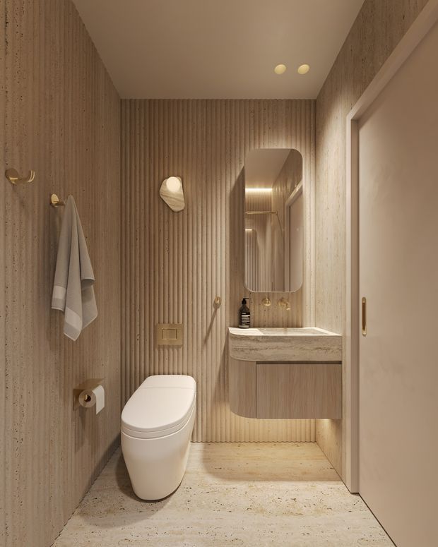 A compact, warm-toned contemporary bathroom with vertically ribbed beige wall finishes, a wall-hung toilet, floating timber-toned vanity, pale stone benchtop, integrated basin, rounded rectangular mirror, and concealed shower area. Soft integrated and wall lighting creates a calm, refined atmosphere.