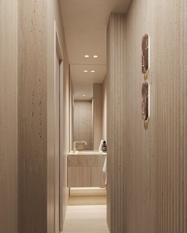 A narrow, softly lit bathroom corridor leading to a compact vanity. The space features continuous warm beige textured wall surfaces, a pale stone benchtop, light timber vanity cabinetry, recessed ceiling lights, and sculptural wall sconces. The partially visible basin and towel confirm the bathroom setting. The design is highly refined, monochromatic, and minimalist.