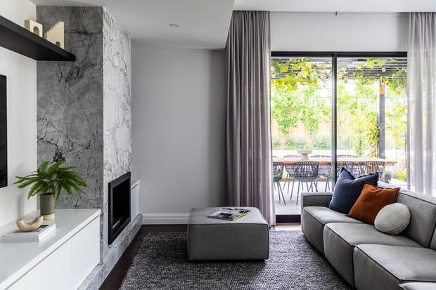 A contemporary living room with a grey sectional sofa, textured dark rug, white built-in media cabinetry, and a dramatic grey marble-clad fireplace wall. Full-height curtains frame large glazed doors opening to an outdoor dining terrace and garden.