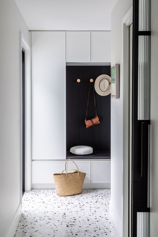 A narrow, bright entryway with built-in white cabinetry, a recessed black vertical-ribbed coat and seating nook, wall hooks, a hat, a small cushion, and a woven basket. The floor is finished in light terrazzo with dark and grey aggregate flecks. A black-framed glazed door or partition is visible at the right edge.