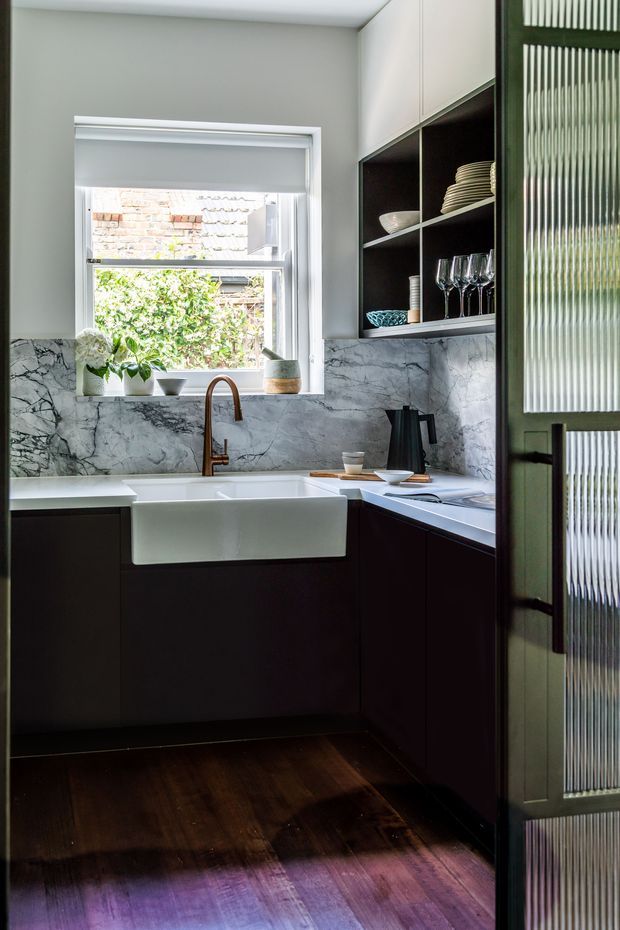 A compact, refined kitchen with dark cabinetry, a deep white farmhouse sink, white marble-look stone splashback, and a copper-toned gooseneck tap. Open shelving displays crockery and glassware, while a ribbed translucent-glass door frames the right side. A window provides natural light above the sink, and dark timber flooring adds warmth.