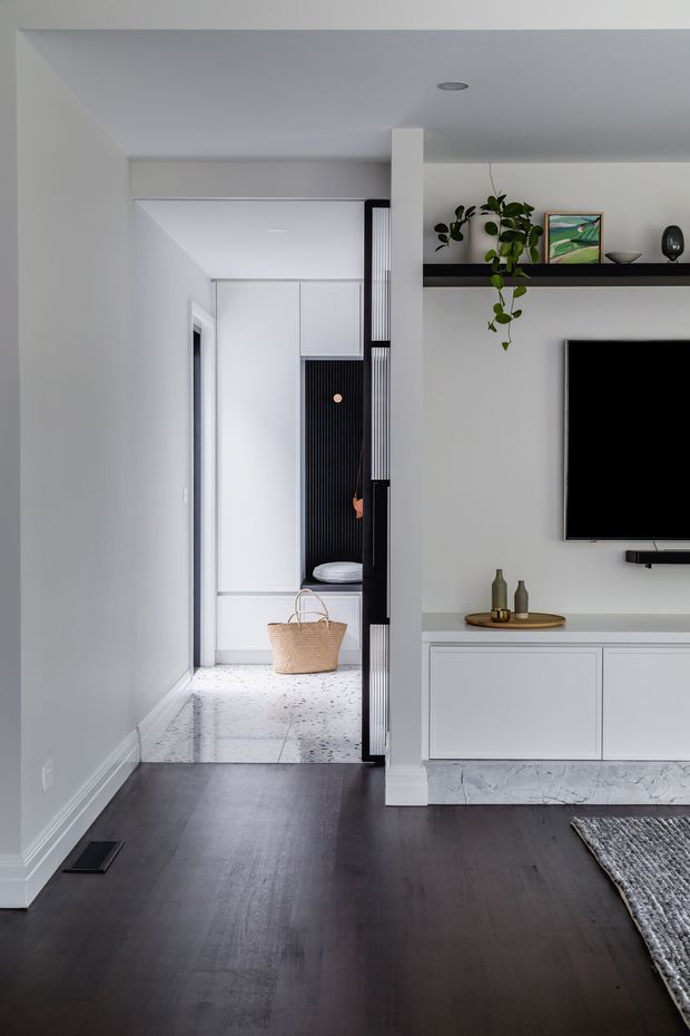 A clean, contemporary open-plan living area viewed toward a hallway and entry zone. The room has dark timber flooring, white painted walls and ceiling, a wall-mounted television above minimalist white built-in cabinetry, and a slim black display shelf with plants and artwork. A central white structural column separates the living area from a bright corridor featuring white joinery, a black vertical slatted accent panel, and pale terrazzo-style flooring. The palette is restrained and monochromatic with natural greenery and warm decorative accents.