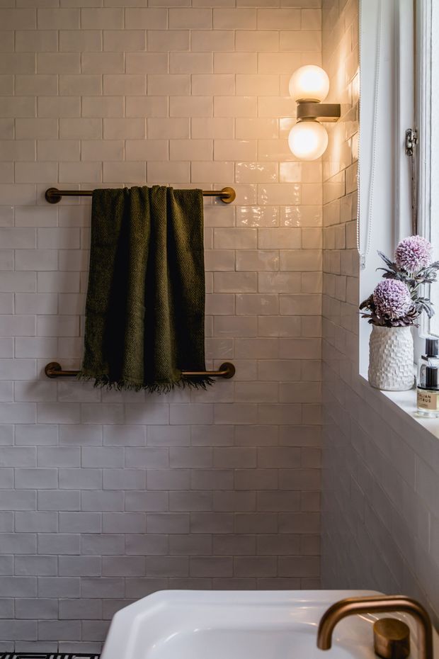 A warmly lit bathroom corner featuring glossy pale pink-beige subway wall tiles, two brass towel rails holding a dark green towel, brass wall sconces with spherical bulbs, and a white freestanding bathtub with a brass tap beneath a window. A textured vase with dried flowers sits on the windowsill.
