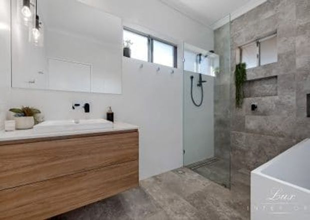 Contemporary bathroom featuring a floating timber vanity with a white benchtop and integrated basin, frameless glass walk-in shower, freestanding white bathtub, black fixtures, and large-format grey stone-look wall and floor tiles. A narrow horizontal window provides natural light, while a recessed wall niche and greenery add detail.