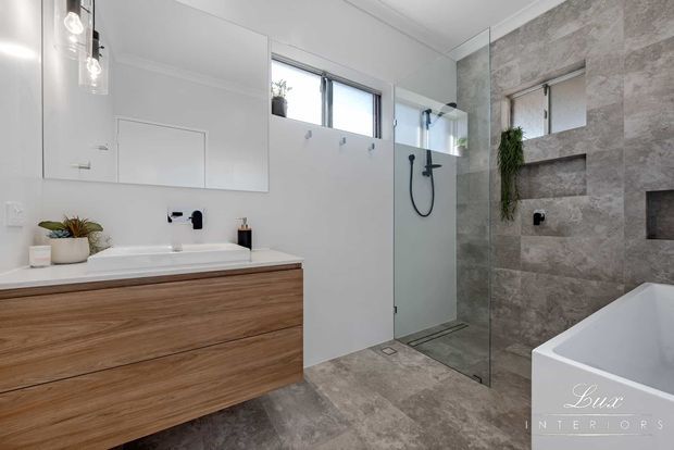 Contemporary bathroom featuring a floating timber vanity with a white benchtop and basin, frameless glass walk-in shower, wall-mounted black fixtures, large-format grey stone-look wall and floor tiles, a freestanding white bathtub, and a mirrored wall cabinet. The room is bright and clean with natural light from a high horizontal window.