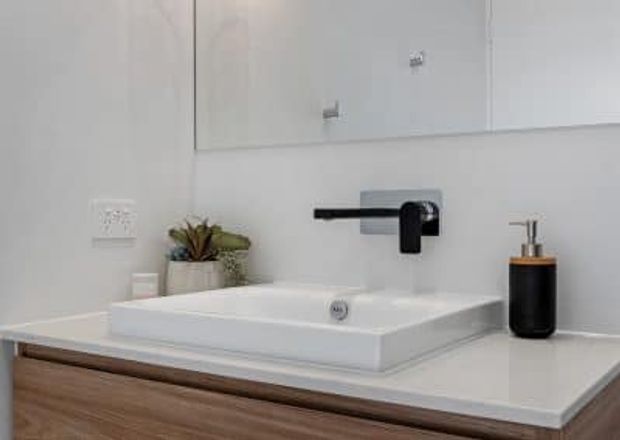 Contemporary bathroom vanity with a white rectangular vessel basin on a light benchtop, timber-look drawer cabinetry, a black wall-mounted tap, and a black soap dispenser. A large mirror and minimal white walls create a clean, refined appearance.