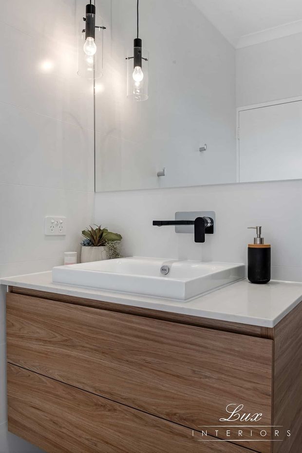 A bright, contemporary bathroom vanity featuring a white vessel basin on a white benchtop, wall-mounted matte black tapware, warm timber-look drawer cabinetry, a large mirrored splashback, and minimalist pendant lights. Small plants and a black soap dispenser add restrained styling.