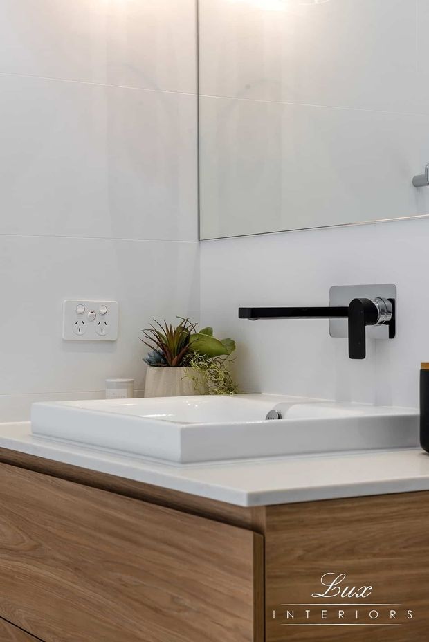 Close-up of a contemporary bathroom vanity featuring a white rectangular countertop basin, wall-mounted matte black tapware, pale large-format wall tiles, a frameless mirror, and warm wood-grain cabinetry. Small potted plants add a natural detail to the clean, refined space.