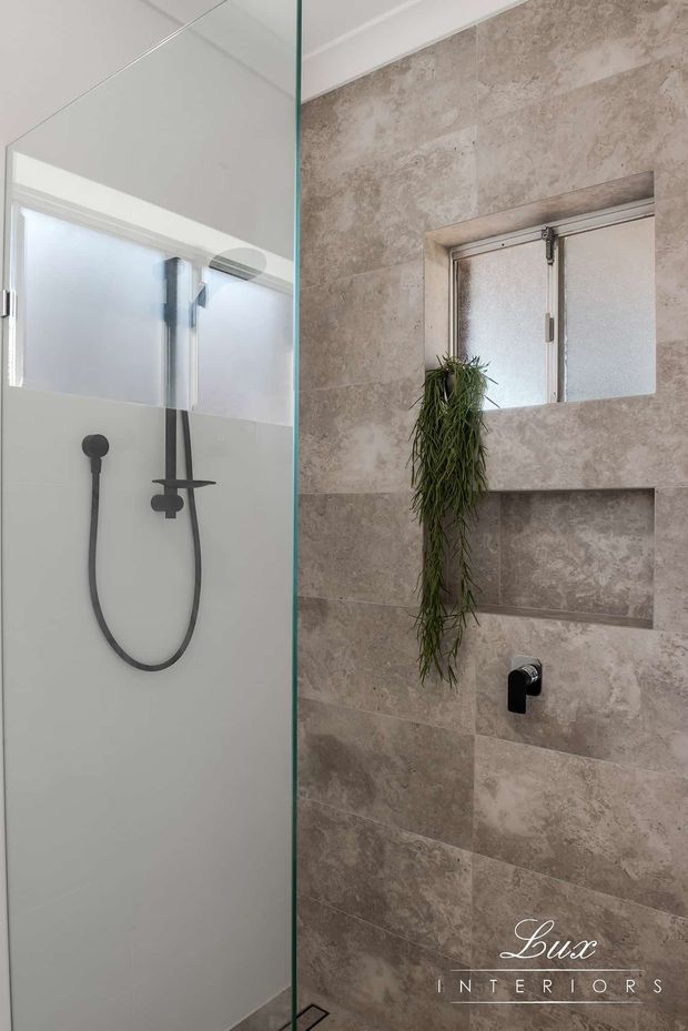 A compact contemporary bathroom shower area viewed through a clear glass partition. The shower features matte black wall-mounted fixtures, a handheld shower rail, pale frosted glazing, and large beige stone-look wall tiles. A recessed wall niche and hanging greenery add detail to the tiled wall.