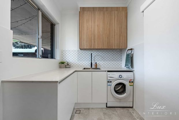 Bright compact laundry room with an L-shaped white benchtop and streamlined white cabinetry. A front-loading washing machine is integrated beneath the bench, with warm timber overhead cabinets and a patterned tiled splashback. The room has a large window, light neutral walls, and pale stone-look floor tiles.
