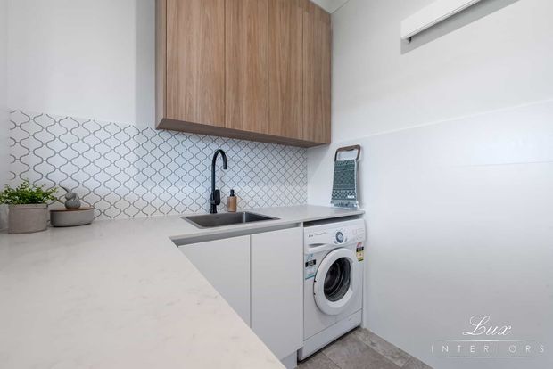 Compact contemporary laundry with white cabinetry, a pale stone benchtop, integrated stainless sink, black gooseneck tap, patterned geometric tile splashback, timber overhead cupboards, and a front-loading washing machine. The room is bright and clean with neutral finishes and warm wood accents.