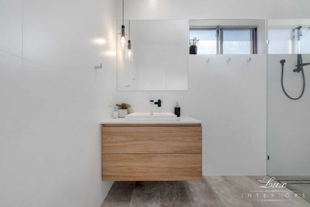 Bright contemporary bathroom with a floating natural timber vanity, white basin and wall-mounted black tapware. Large white wall tiles, a mirrored cabinet, pendant lights, a high horizontal window, and a frameless glass shower with black fixtures create a clean, minimal interior.