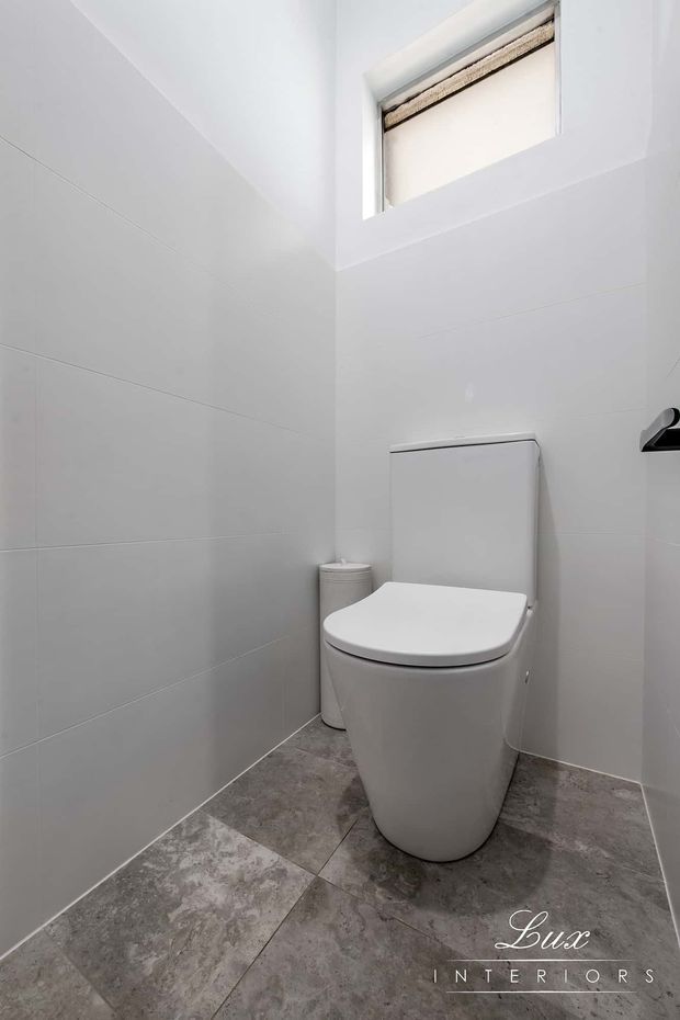 Compact modern toilet room with a wall-mounted cistern and contemporary white toilet, white tiled walls, and grey stone-look floor tiles. A high frosted or roller-blind-covered window provides natural light, while a small black wall-mounted fixture is visible at right.