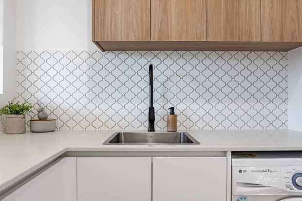 Close-up of a compact contemporary kitchen with white cabinetry and benchtops, a stainless-steel undermount sink, matte black gooseneck tap, pale timber overhead cabinets, and a decorative white-and-grey geometric tiled splashback. A front-loading washing machine is integrated beneath the benchtop at right.