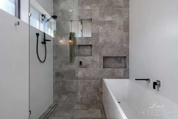 Contemporary minimalist bathroom with a walk-in glass shower, freestanding white bathtub, black fixtures, and large grey stone-look wall and floor tiles. Recessed wall niches and a narrow high window add practical detail and natural light.
