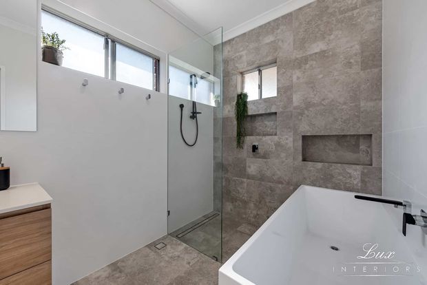 Contemporary bathroom featuring a frameless glass walk-in shower, freestanding white bathtub, wall-mounted black fixtures, a textured stone-look feature wall with recessed niches, pale tiled walls and floor, and timber-toned vanity cabinetry. High horizontal windows provide natural light.