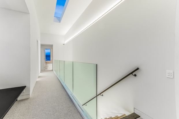 A bright, minimalist upper-level hallway or landing overlooking a stairwell. White walls and ceiling are accented by a long linear recessed light and a narrow skylight, while a frosted glass balustrade with a dark metal handrail lines the stairs. Light carpet and pale timber stair treads create a clean, contemporary feel.