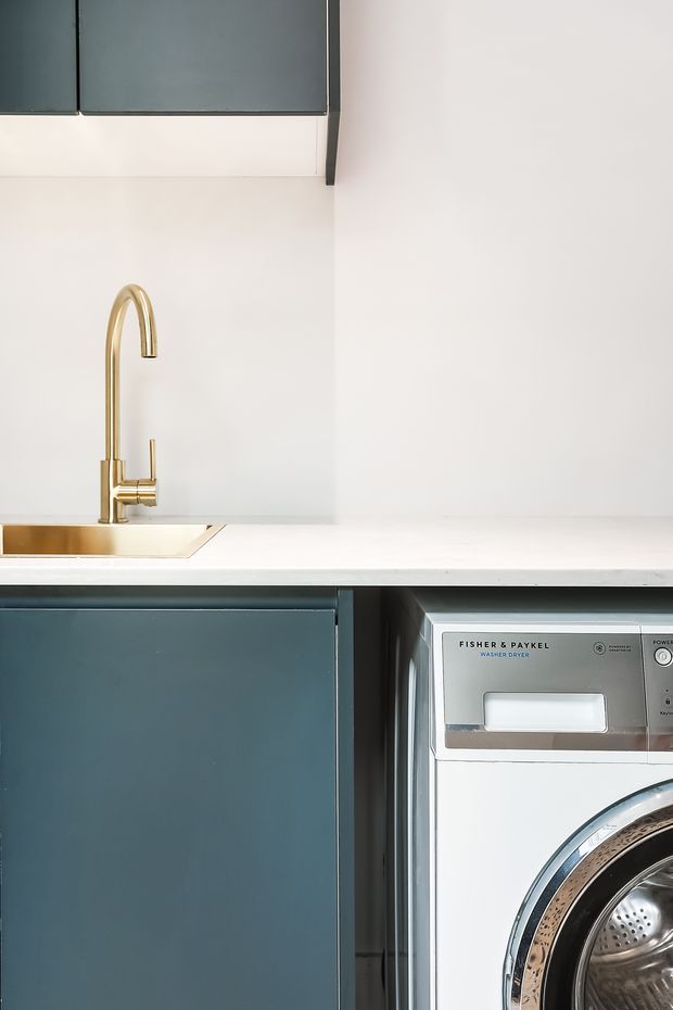 Close-up view of a compact laundry area with deep blue-green flat-panel cabinetry, a white benchtop and splashback, and a brass-gold sink with a tall curved mixer tap. A front-loading Fisher & Paykel washer-dryer is integrated beneath the benchtop at right. The composition is clean and contemporary with minimal visible decoration.