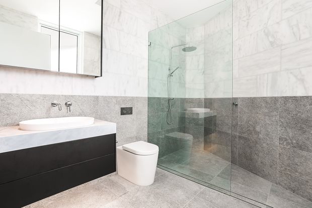 Bright contemporary bathroom featuring a wall-mounted black vanity with a white vessel basin, wall-mounted tapware, a white toilet, and a frameless glass walk-in shower. Large white marble-look wall tiles contrast with grey stone-look tiles on the lower walls and floor. A mirrored cabinet and small window add light and functionality.