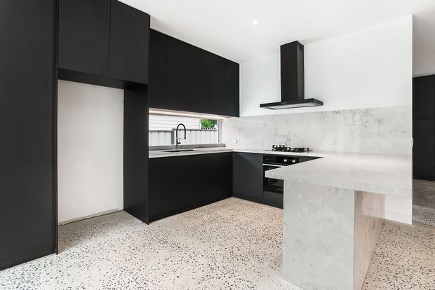 Contemporary minimalist kitchen with matte black full-height cabinetry, a white marble-look splashback and waterfall island, integrated appliances, and a terrazzo-style floor. A window above the sink brings in natural light.