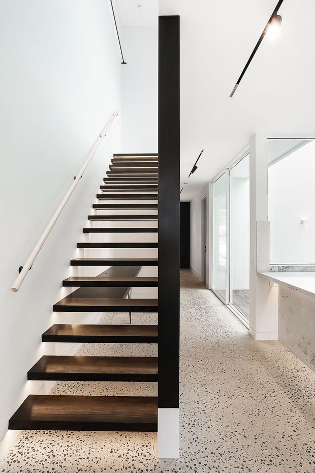 Bright minimalist interior showing an open-riser staircase with dark timber treads, a slender pale handrail, and a black structural spine. The stair rises beside a narrow circulation area with full-height glazed doors and a partially visible white kitchen counter. The floor is light terrazzo with dark aggregate, and linear black ceiling lights accent the clean white interior.