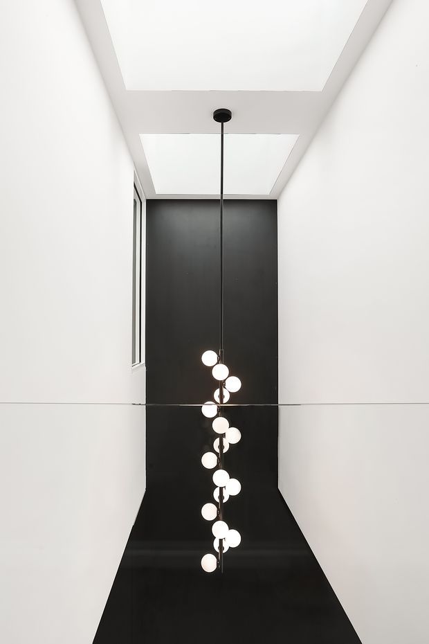 A minimalist vertical interior view of a narrow, double-height space. Crisp white walls and ceiling frame a dramatic black feature wall and black floor, with two large rectangular skylights above. A long black pendant suspends a sculptural cluster of glowing spherical bulbs through the void. A slim window is visible on the left wall.