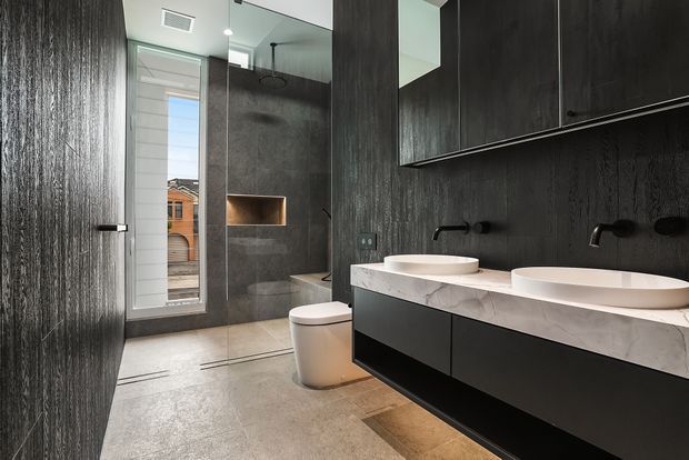 Contemporary double-vanity bathroom with dark textured wall finishes, large mirrored cabinetry, a pale stone benchtop, two vessel basins, matte black tapware, a wall-hung toilet and a glass-enclosed shower. A narrow window provides daylight and an urban view.