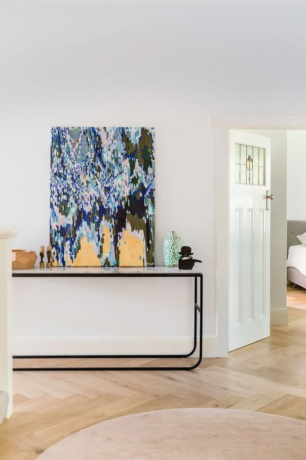 Bright, refined residential entry or hallway with pale painted walls, light herringbone timber flooring, and a slim black metal console table. A large abstract blue, green, cream and ochre artwork hangs above the console, accompanied by decorative vessels. An open paneled door with leaded glass provides a view into a bedroom, while a soft neutral rug occupies the foreground.