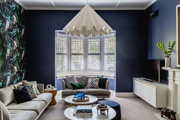 A refined living room with deep navy walls, a pale upholstered sectional and armchair, layered patterned cushions, a sculptural scalloped pendant light, and a white bay window fitted with plantation shutters. A white fireplace surround and low media cabinet frame the room, with round coffee tables and dark wood accents completing the space.