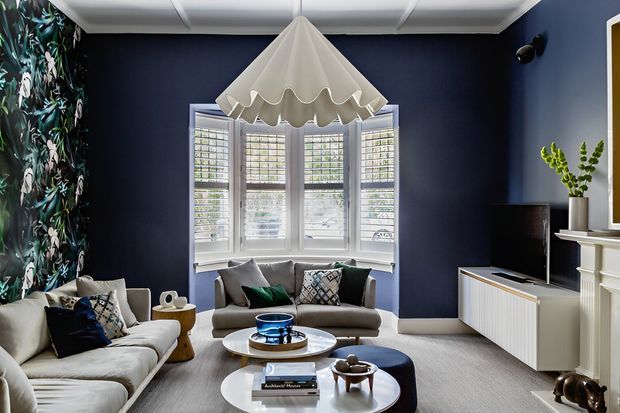 A refined living room with deep navy walls, a white bay window fitted with plantation shutters, pale upholstered sofas, a round coffee table, a sculptural pendant light, and white built-in cabinetry. Botanical wallpaper forms a feature wall at left, with layered cushions and decor adding pattern and texture.
